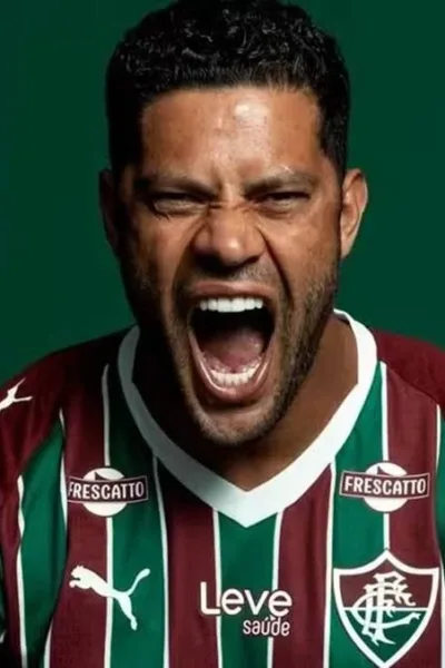 Atacante Hulk em imagem, horizontal, de anúncio de sua vinda para o Fluminense a partir de 2026. Ele está com a camisa do time e dá um grito. Escudo do time está acima de seu nome, à esquerda do fundo verde, gravado em branco.