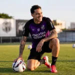 Jogador do Ceará FC ajoelhado apoiado sobre um dos joelhos, sorrindo, olhando de canto, fim de tarde, segurando bola com mão direita. Imagem gerada por IA.