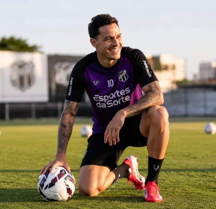 Jogador do Ceará FC ajoelhado apoiado sobre um dos joelhos, sorrindo, olhando de canto, fim de tarde, segurando bola com mão direita. Imagem gerada por IA.