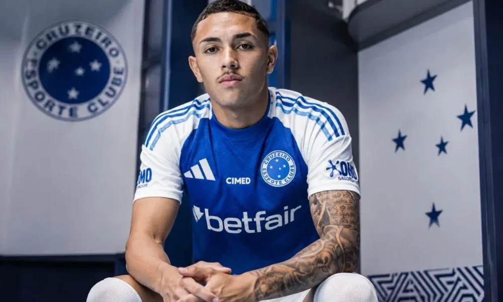 Kauã Moraes, do Cruzeiro, sentado em vestiário com mãos cruzadas e braços sobre os joelhos, expressão neutra. Imagem gerada por IA.