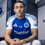 Kauã Moraes, do Cruzeiro, sentado em vestiário com mãos cruzadas e braços sobre os joelhos, expressão neutra. Imagem gerada por IA.