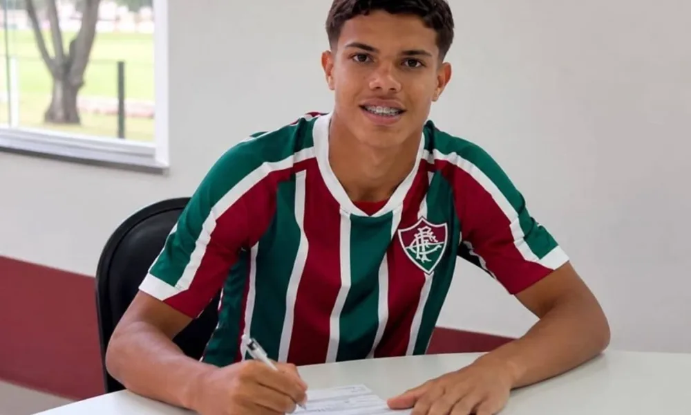 Julio Fidelis assinando renovação com Fluminense. Jogador está com camisa do time, braços sobre mesa branca com papel e caneta, sorrindo para câmera posando para foto.