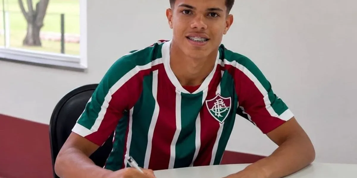 Julio Fidelis assinando renovação com Fluminense. Jogador está com camisa do time, braços sobre mesa branca com papel e caneta, sorrindo para câmera posando para foto.
