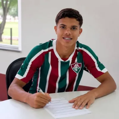 Julio Fidelis assinando renovação com Fluminense. Jogador está com camisa do time, braços sobre mesa branca com papel e caneta, sorrindo para câmera posando para foto.