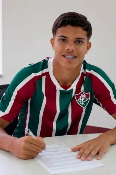 Fluminense Amplia Vínculo de Júlio Fidelis Até 2031 Julio Fidelis assinando renovação com Fluminense. Jogador está com camisa do time, braços sobre mesa branca com papel e caneta, sorrindo para câmera posando para foto.