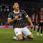 Kevin Serna, de joelhos comemorando, apontando para o escudo do time, em partida contra o Vitória, empatando em 2x2. Imagem gerada por IA.