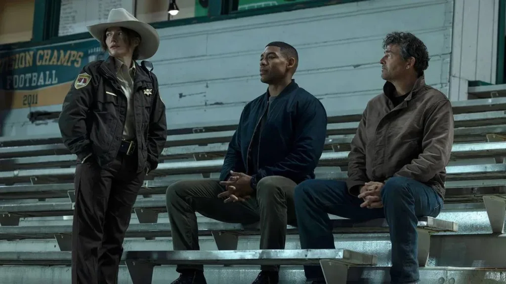Aaron Pierre (esquerda) e Kyle Chandler (direita) como dois lanternas na nova série adaptada dos quadrinhos da DC sobre os heróis, interpretando John Stewart e Hal Jordan, respectivamente. Ambos estão sentados frente à xerife Kerry (Kelly Macdonald) à esquerda, em uma arquibancada.