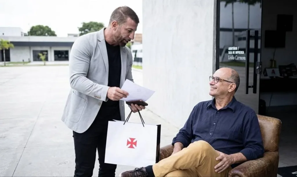 Vasco Eterniza Ledio Carmona no CT Moacyr Barbosa em Homenagem Emocionante 2 Homenagem a Ledio Carmona no CT do Vasco. Ele está sentado em uma cadeira, sorrindo, recebe um presente do time.