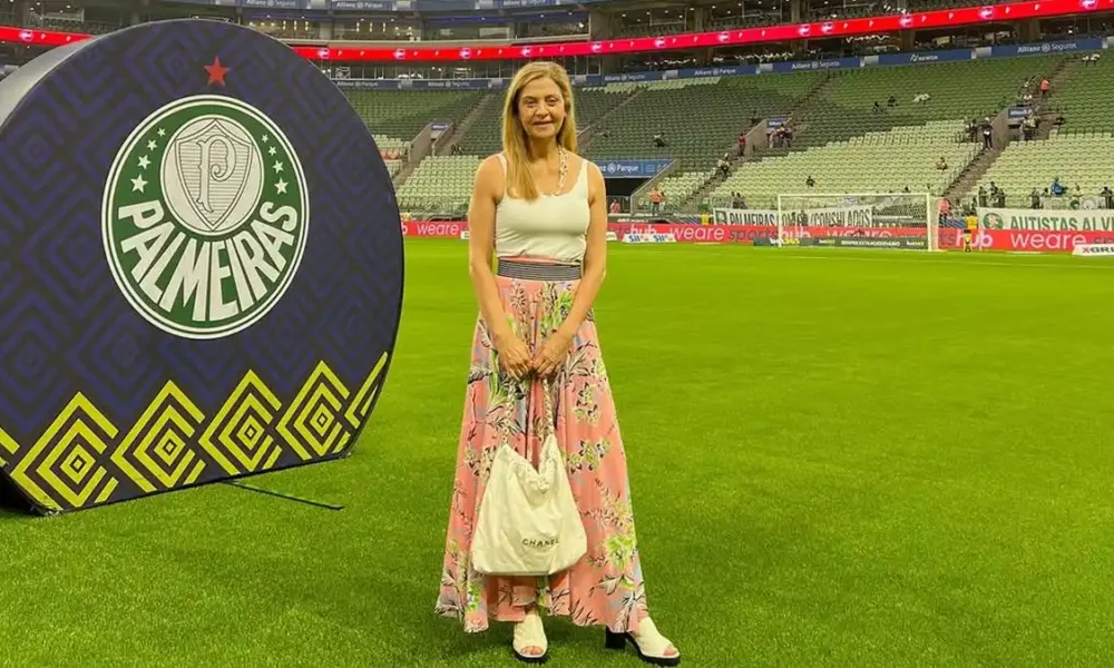 Leila Pereira, presidente do Palmeiras, na Arena Palmeiras em março de 2026. Ela está de pé no campo com um vestido e posando sorrindo segurando uma bolsa.