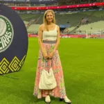Leila Pereira, presidente do Palmeiras, na Arena Palmeiras em março de 2026. Ela está de pé no campo com um vestido e posando sorrindo segurando uma bolsa.