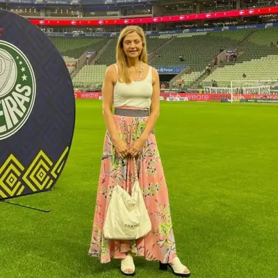 Leila Pereira, presidente do Palmeiras, na Arena Palmeiras em março de 2026. Ela está de pé no campo com um vestido e posando sorrindo segurando uma bolsa.