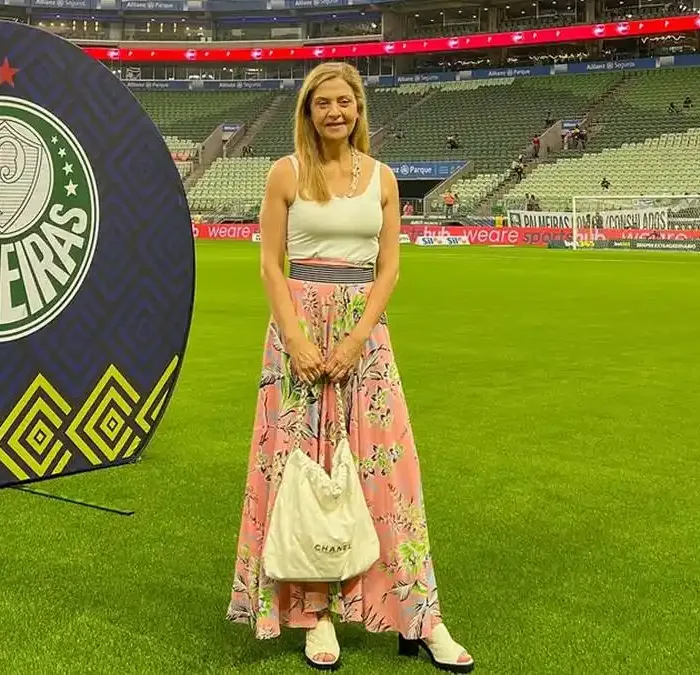 Leila Pereira, presidente do Palmeiras, na Arena Palmeiras em março de 2026. Ela está de pé no campo com um vestido e posando sorrindo segurando uma bolsa.