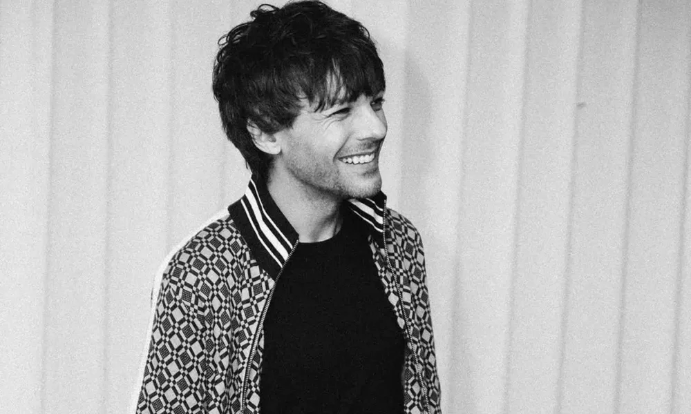 Louis Tomlinson, ligeiramente de corpo virado, sorriso no rosto, foto em preto e branco com persianas brancas ao fundo.