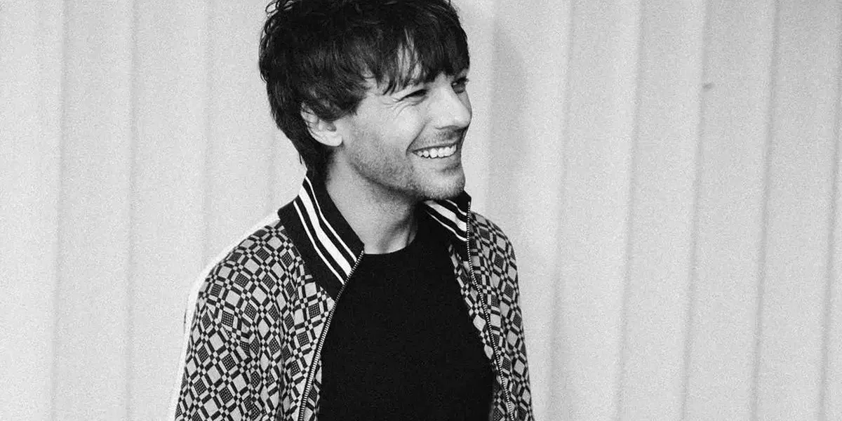 Louis Tomlinson, ligeiramente de corpo virado, sorriso no rosto, foto em preto e branco com persianas brancas ao fundo.