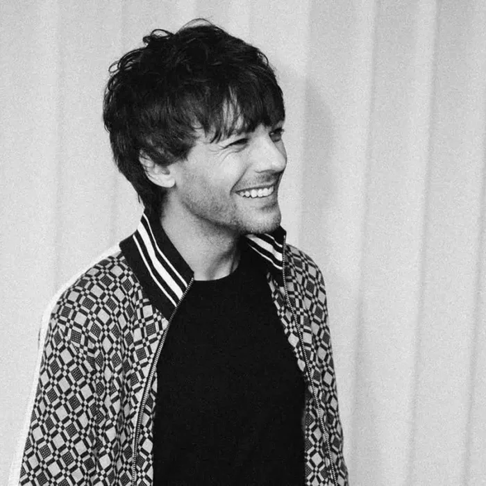 Louis Tomlinson, ligeiramente de corpo virado, sorriso no rosto, foto em preto e branco com persianas brancas ao fundo.
