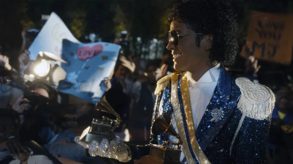 Jaafar Jackson caracterizado como seu tio, Michael Jackson, no filme biográfico "Michael", ele está com uma roupa brilhante azul de baile com ombreira, óculos escuros, segurando dois prêmios Grammy.
