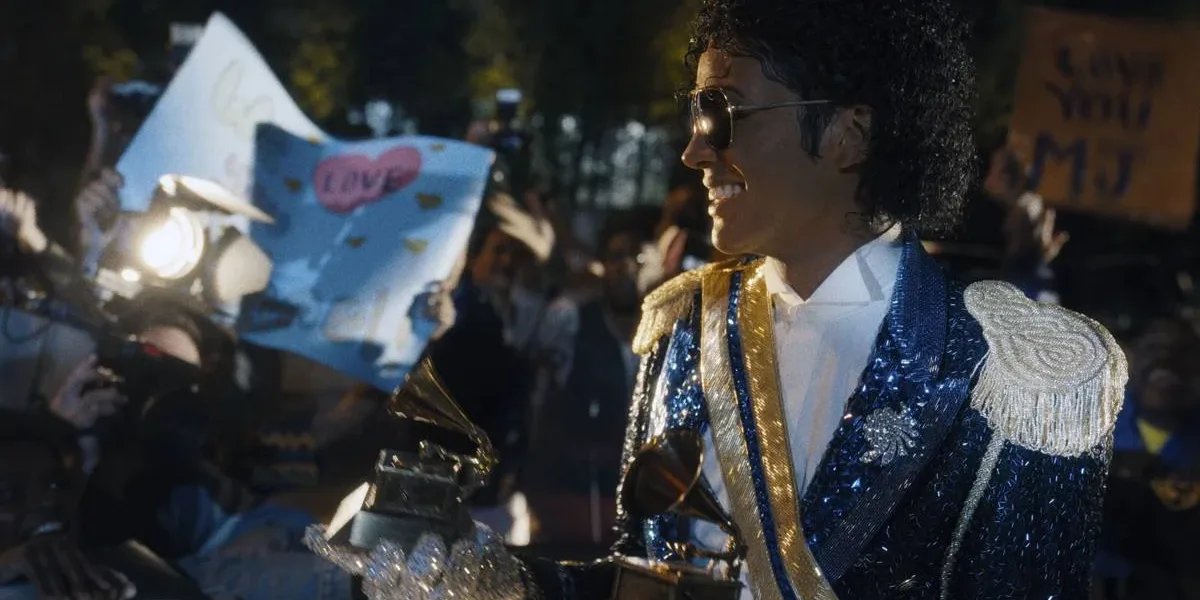 Michael 2 | Sequência da Cinebiografia Enfrenta Desafios para Retratar Fase mais Controversa de Michael Jackson Jaafar Jackson caracterizado como seu tio, Michael Jackson, no filme biográfico "Michael", ele está com uma roupa brilhante azul de baile com ombreira, óculos escuros, segurando dois prêmios Grammy.