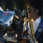 Jaafar Jackson caracterizado como seu tio, Michael Jackson, no filme biográfico "Michael", ele está com uma roupa brilhante azul de baile com ombreira, óculos escuros, segurando dois prêmios Grammy.