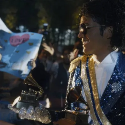 Jaafar Jackson caracterizado como seu tio, Michael Jackson, no filme biográfico "Michael", ele está com uma roupa brilhante azul de baile com ombreira, óculos escuros, segurando dois prêmios Grammy.