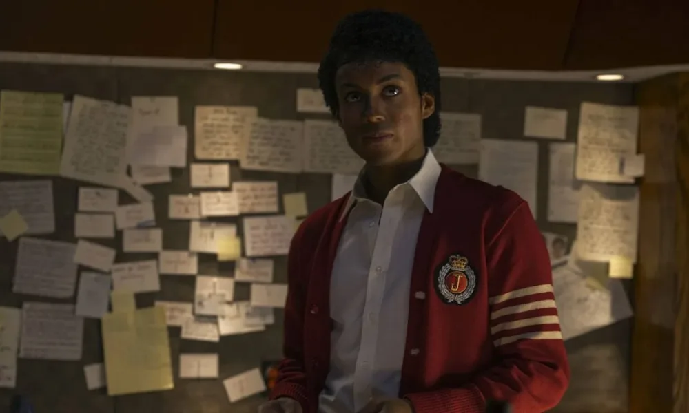 Jaafar Jackson caracterizado como seu tio, Michael Jackson, no filme biográfico "Michael"; ele está com um semblante sério, blusa colegial vermelha, olhando levemente para cima com um quadro cheio de pins atrás.