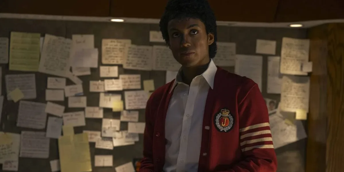 Jaafar Jackson caracterizado como seu tio, Michael Jackson, no filme biográfico "Michael"; ele está com um semblante sério, blusa colegial vermelha, olhando levemente para cima com um quadro cheio de pins atrás.