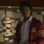 Jaafar Jackson caracterizado como seu tio, Michael Jackson, no filme biográfico "Michael"; ele está com um semblante sério, blusa colegial vermelha, olhando levemente para cima com um quadro cheio de pins atrás.
