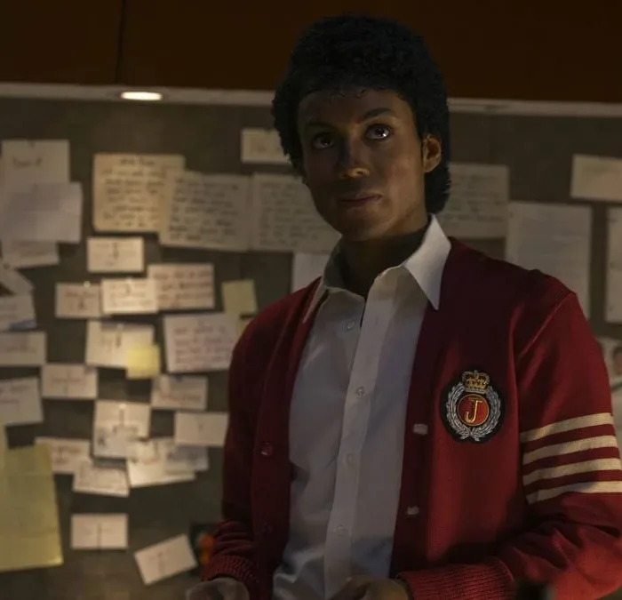 Jaafar Jackson caracterizado como seu tio, Michael Jackson, no filme biográfico "Michael"; ele está com um semblante sério, blusa colegial vermelha, olhando levemente para cima com um quadro cheio de pins atrás.