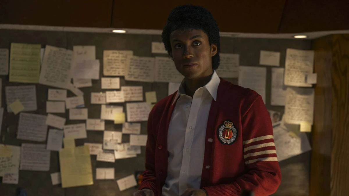 Jaafar Jackson caracterizado como seu tio, Michael Jackson, no filme biográfico "Michael"; ele está com um semblante sério, blusa colegial vermelha, olhando levemente para cima com um quadro cheio de pins atrás.