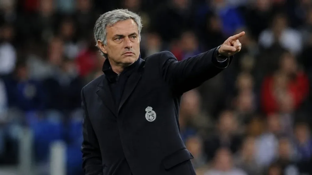 Real Madrid e Mourinho: Dois Gigantes Ligados Pelo Destino 1 Treinador José Mourinho, braço esquerdo levantado e apontando o dedo, expressão séria, em partida de futebol, fundo da torcida embaçado. Ele está com escudo do Real Madrid em jaqueta técnica. Imagem gerada por IA.
