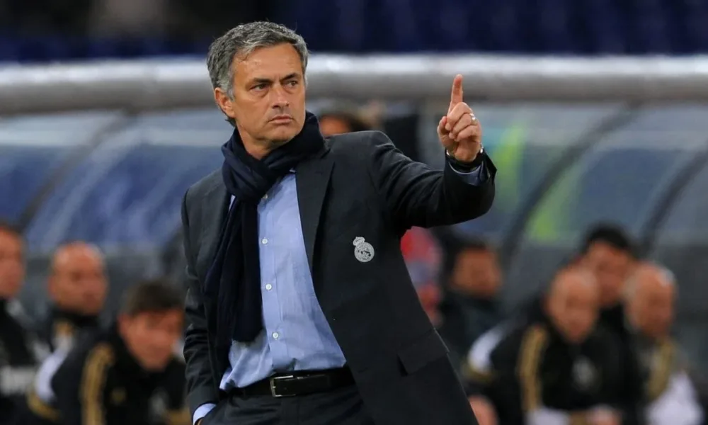 Treinador José Mourinho, braço esquerdo levantado e apontando o dedo para cima, expressão séria, em partida de futebol, fundo da torcida embaçado com destaque para banco de reservas. Ele está com escudo do Real Madrid em jaqueta técnica, aberta. Imagem gerada por IA.