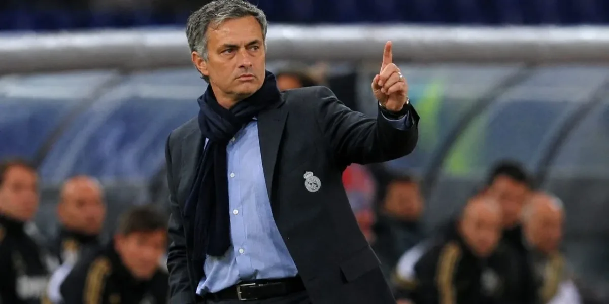 Real Madrid e Mourinho: Dois Gigantes Ligados Pelo Destino Treinador José Mourinho, braço esquerdo levantado e apontando o dedo para cima, expressão séria, em partida de futebol, fundo da torcida embaçado com destaque para banco de reservas. Ele está com escudo do Real Madrid em jaqueta técnica, aberta. Imagem gerada por IA.