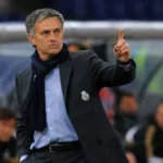 Treinador José Mourinho, braço esquerdo levantado e apontando o dedo para cima, expressão séria, em partida de futebol, fundo da torcida embaçado com destaque para banco de reservas. Ele está com escudo do Real Madrid em jaqueta técnica, aberta. Imagem gerada por IA.