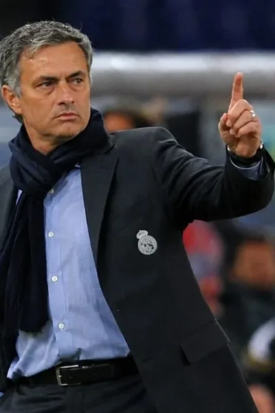 Treinador José Mourinho, braço esquerdo levantado e apontando o dedo para cima, expressão séria, em partida de futebol, fundo da torcida embaçado com destaque para banco de reservas. Ele está com escudo do Real Madrid em jaqueta técnica, aberta. Imagem gerada por IA.