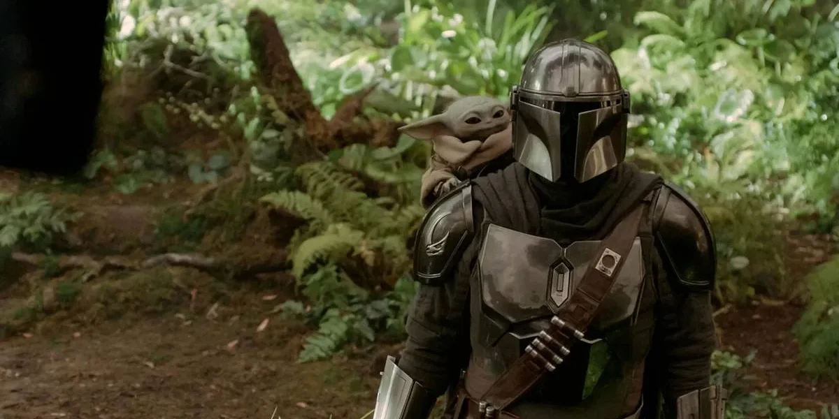 O Mandaloriano e Grogu | Filme Resgata a Essência Clássica de Star Wars nos Cinemas O Mandaloriano, na metade direita da tela, em uma paisagem de floresta, com Grogu no ombro. Imagem de "O Mandaloriano e Grogu".