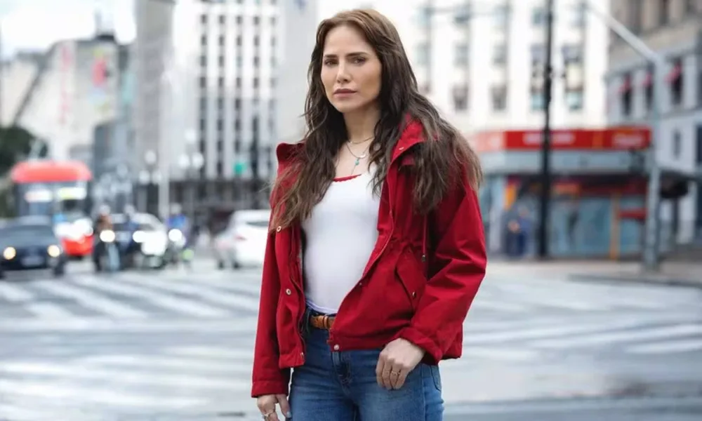Letícia Collin como personagem protagonista da nova novela das 9, "Quem Ama Cuida". Ela está de jaqueta vermelha e uma paisagem urbana de São Paulo ao fundo.