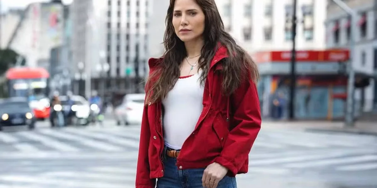Letícia Collin como personagem protagonista da nova novela das 9, "Quem Ama Cuida". Ela está de jaqueta vermelha e uma paisagem urbana de São Paulo ao fundo.