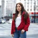 Letícia Collin como personagem protagonista da nova novela das 9, "Quem Ama Cuida". Ela está de jaqueta vermelha e uma paisagem urbana de São Paulo ao fundo.