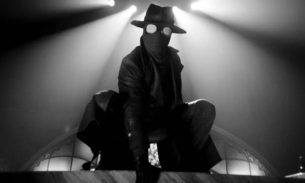 Homem Aranha Noir em série homônima da Prime Video, vestido o traje preto, sobretudo, e um chapéu de investigador em imagem em preto e branco no estilo de filmes policíacos dos anos 30 e 40; ele está agachado como um sapo sobre uma superfície plana. Luzes acima dele ao fundo.