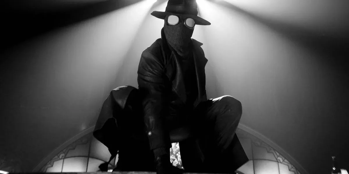 Homem Aranha Noir em série homônima da Prime Video, vestido o traje preto, sobretudo, e um chapéu de investigador em imagem em preto e branco no estilo de filmes policíacos dos anos 30 e 40; ele está agachado como um sapo sobre uma superfície plana. Luzes acima dele ao fundo.