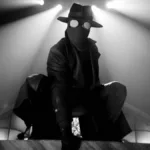 Homem Aranha Noir em série homônima da Prime Video, vestido o traje preto, sobretudo, e um chapéu de investigador em imagem em preto e branco no estilo de filmes policíacos dos anos 30 e 40; ele está agachado como um sapo sobre uma superfície plana. Luzes acima dele ao fundo.
