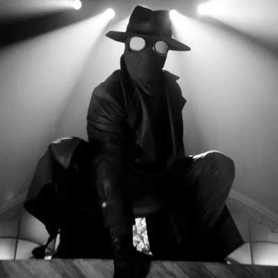 Homem Aranha Noir em série homônima da Prime Video, vestido o traje preto, sobretudo, e um chapéu de investigador em imagem em preto e branco no estilo de filmes policíacos dos anos 30 e 40; ele está agachado como um sapo sobre uma superfície plana. Luzes acima dele ao fundo.