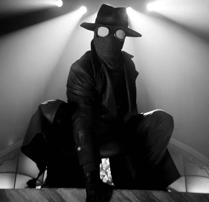 Homem Aranha Noir em série homônima da Prime Video, vestido o traje preto, sobretudo, e um chapéu de investigador em imagem em preto e branco no estilo de filmes policíacos dos anos 30 e 40; ele está agachado como um sapo sobre uma superfície plana. Luzes acima dele ao fundo.