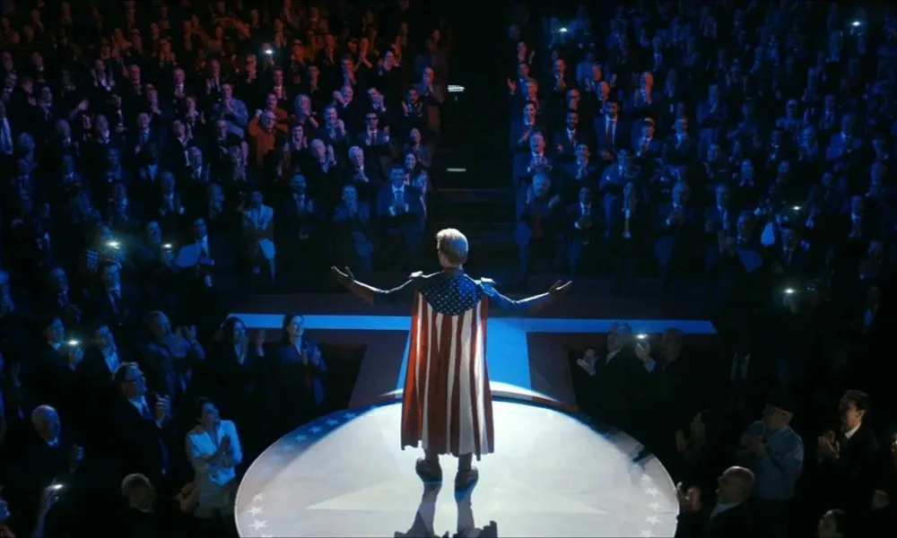 Homelander em um ambiente escuro iluminado apenas no palco, com ele em destaque, braços abertos, uma multidão em um nível abaixo, majoritariamente masculina, aplaudindo eufórica. Imagem do 1º episódio da 5ª temporada de "The Boys".