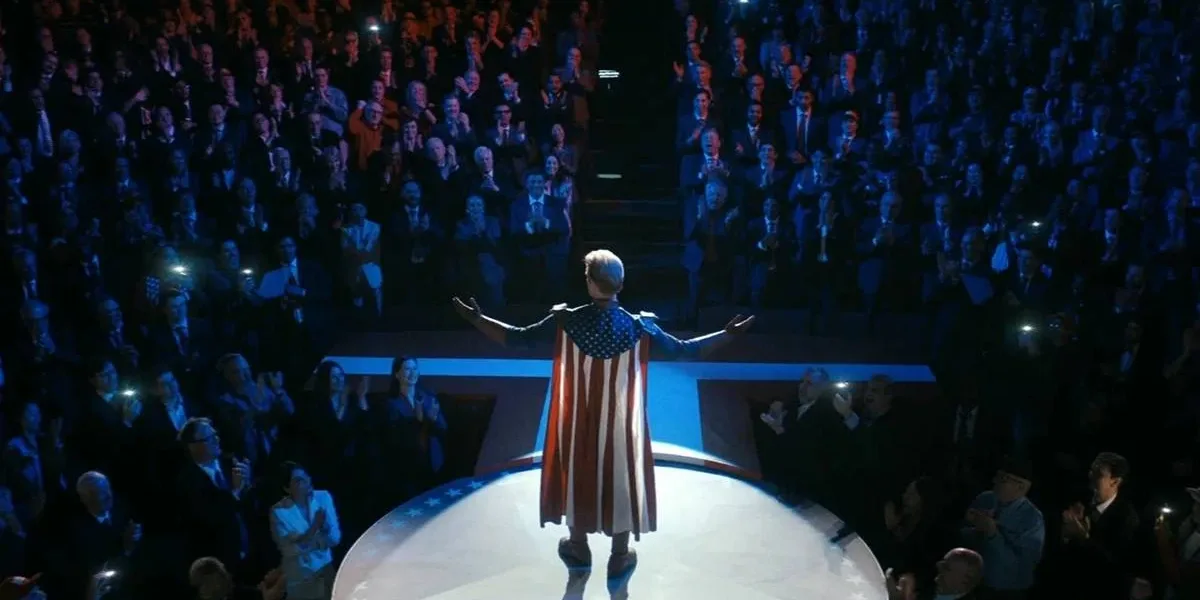 The Boys – 5ª Temporada | Todo Carnaval Tem Seu Fim Homelander em um ambiente escuro iluminado apenas no palco, com ele em destaque, braços abertos, uma multidão em um nível abaixo, majoritariamente masculina, aplaudindo eufórica. Imagem do 1º episódio da 5ª temporada de "The Boys".
