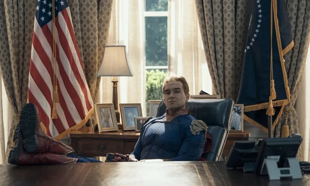 Antony Starr como o Homelander no 7º episódio da 5ª temporada de The Boys. Ele está com os pés sobre a mesa, bandeira dos EUA ao fundo, uma expressão inquieta.