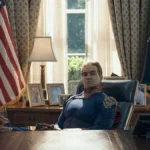 Antony Starr como o Homelander no 7º episódio da 5ª temporada de The Boys. Ele está com os pés sobre a mesa, bandeira dos EUA ao fundo, uma expressão inquieta.