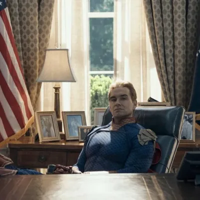 Antony Starr como o Homelander no 7º episódio da 5ª temporada de The Boys. Ele está com os pés sobre a mesa, bandeira dos EUA ao fundo, uma expressão inquieta.