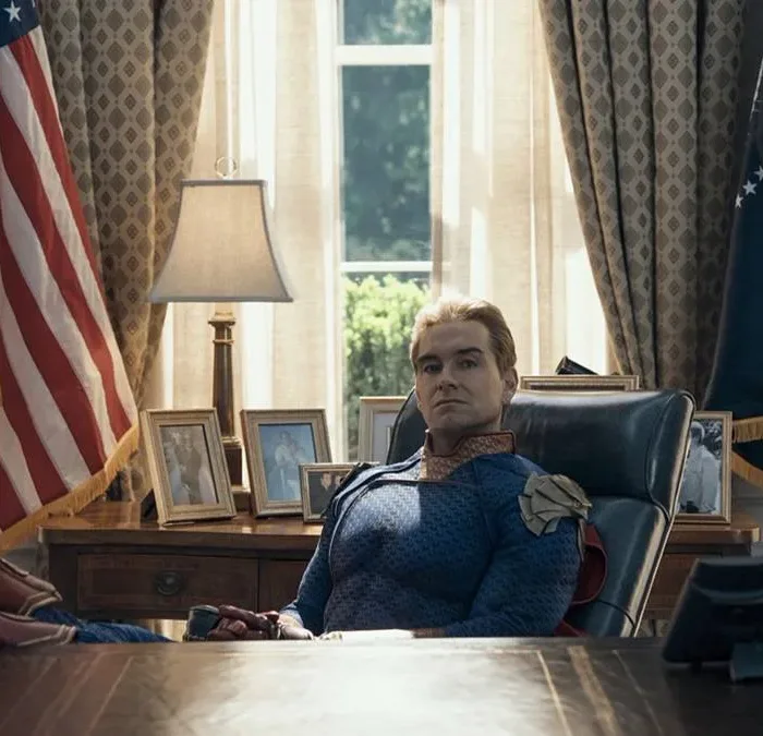 Antony Starr como o Homelander no 7º episódio da 5ª temporada de The Boys. Ele está com os pés sobre a mesa, bandeira dos EUA ao fundo, uma expressão inquieta.