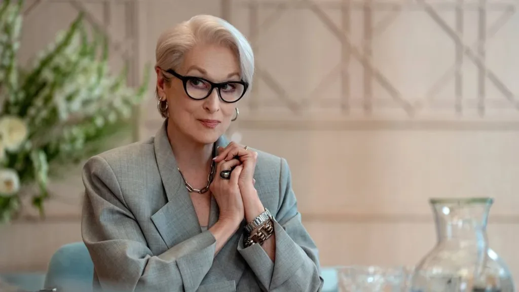 Meryl Streep como Miranda Priestly, mãos trançadas juntas ao peito, vestida sentada com um sorriso em cadeira na empresa Runaway.