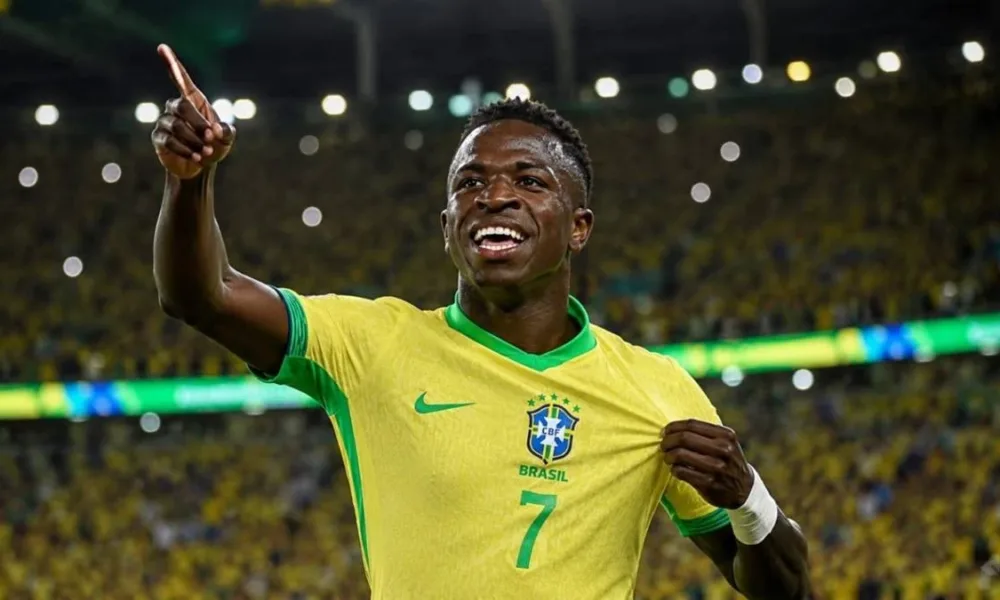 Vini Jr. levanta o braço direito e puxa camisa da seleção, comemorando em partida, sorrindo. Imagem gerada por IA.